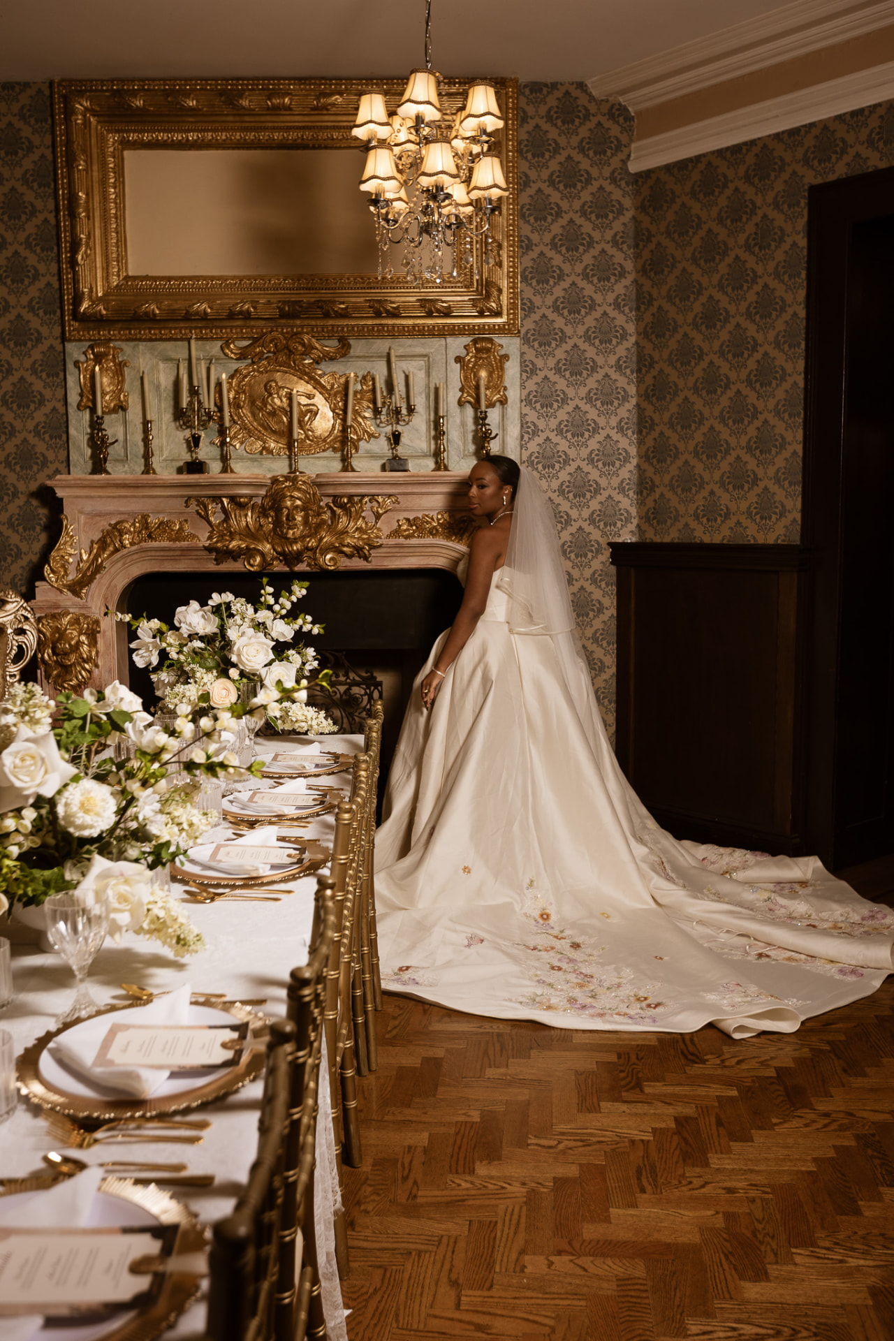 Bride in a floral-embellished gown beside an ornate fireplace and reception table